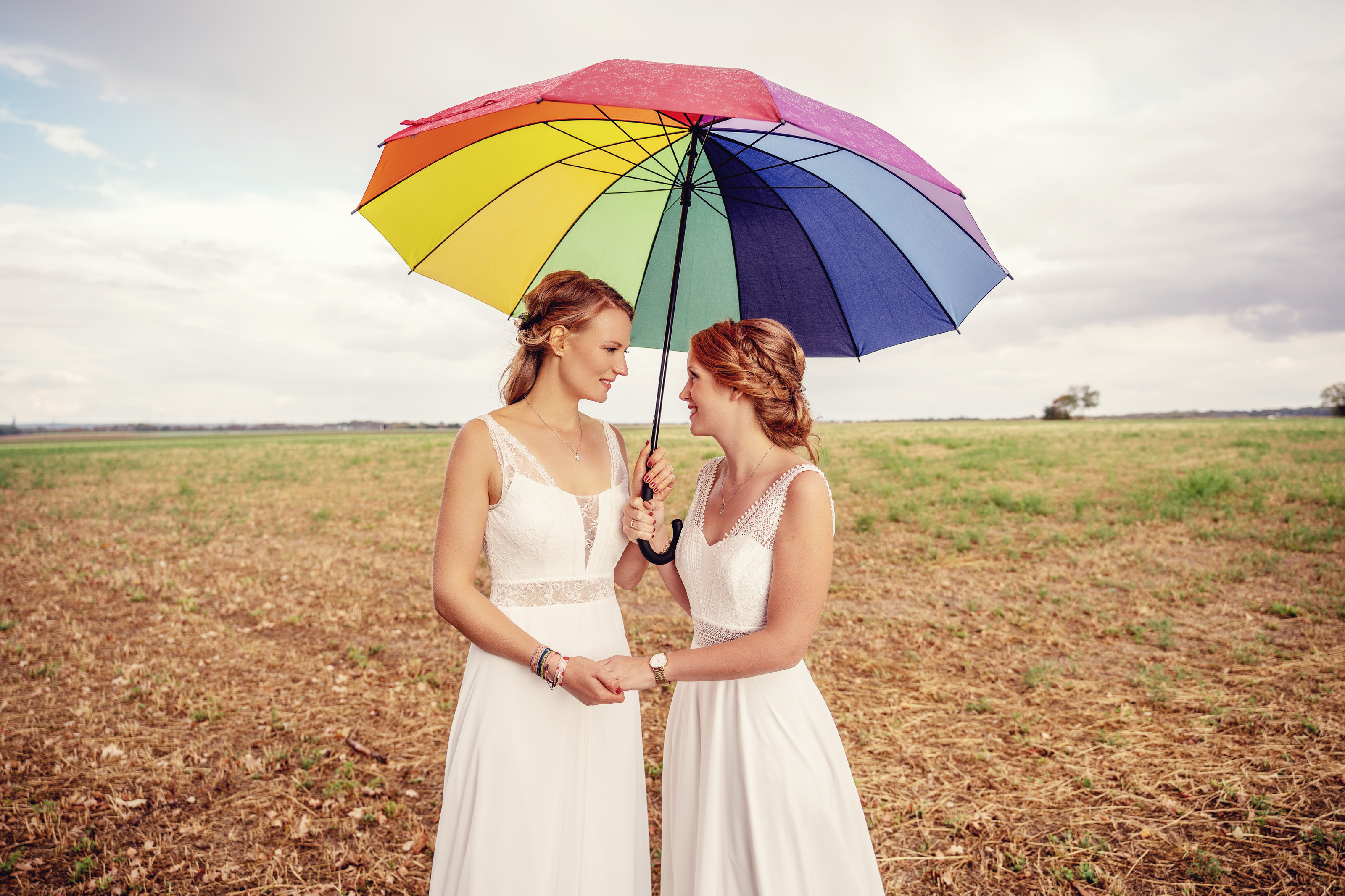 Hochzeitsfotograf Bielefeld – Zwei Bräute auf weitem Feld mit Regenbogenschirm, lachen sich an, bewölkter Himmel
