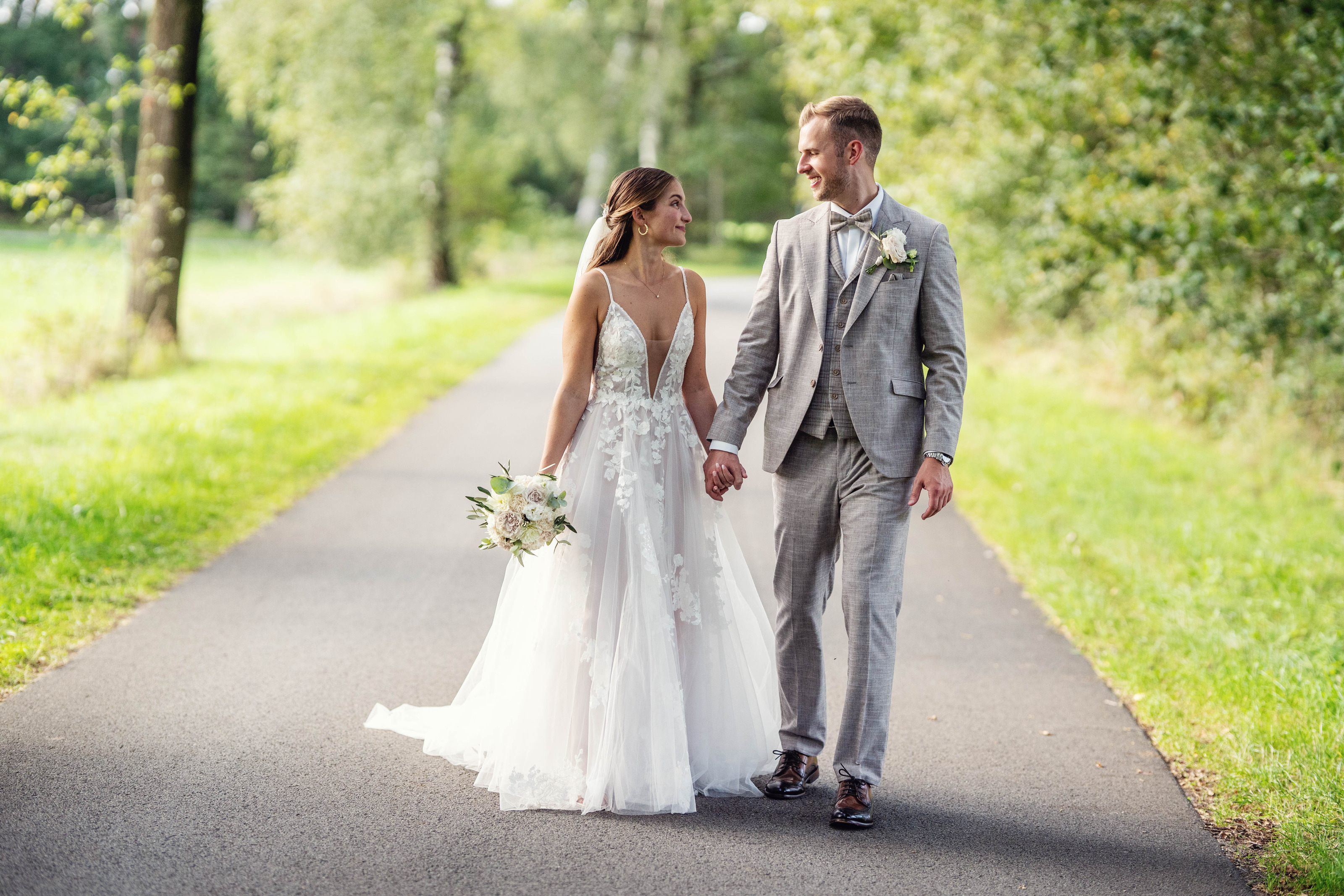 Hochzeitsfotograf Bielefeld – Brautpaar spaziert Hand in Hand auf baumgesäumter Allee, schauen sich verliebt an