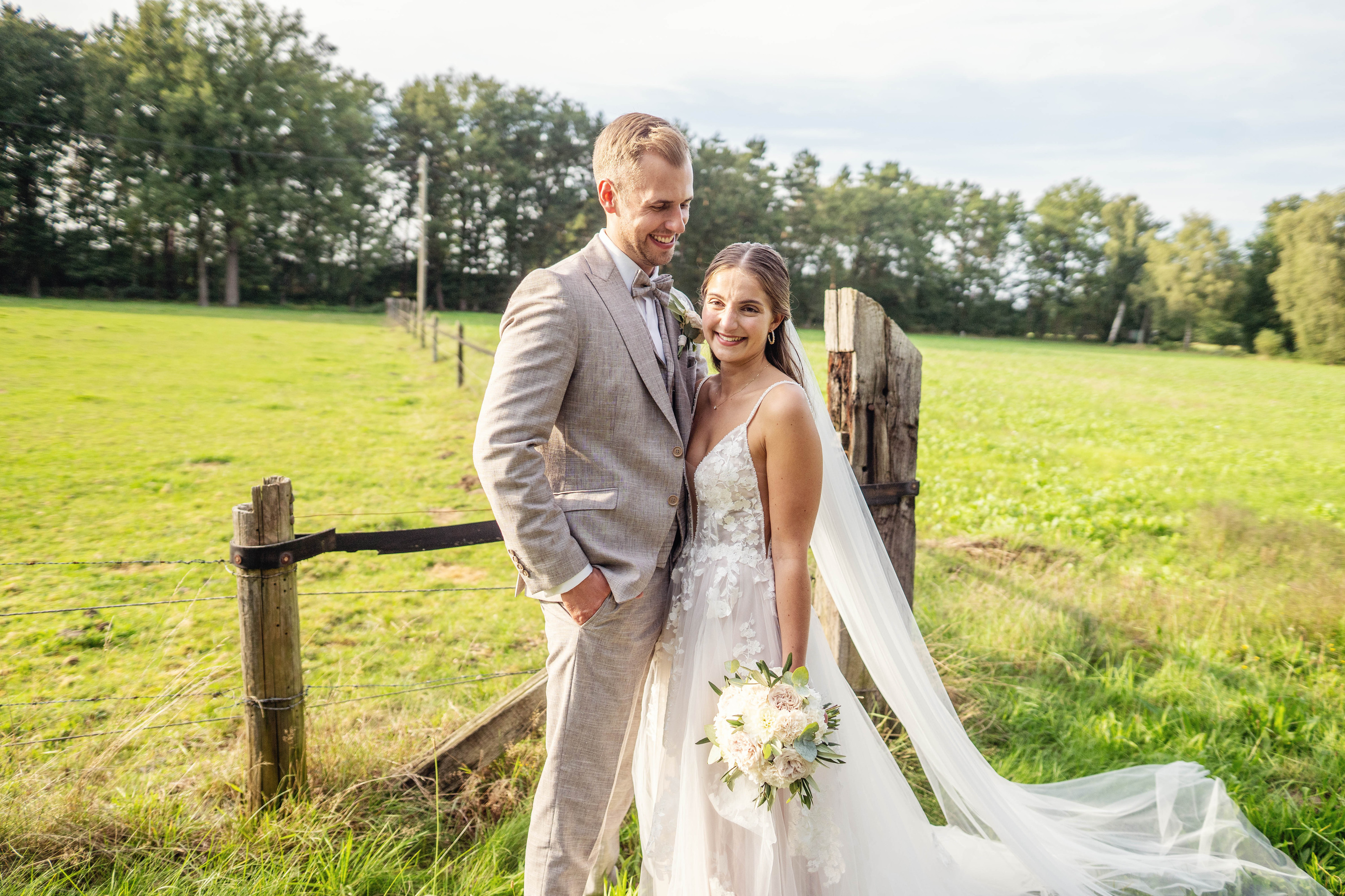 Hochzeitsfotograf Bielefeld – Brautpaar steht lächelnd an Holzzaun auf sonniger Wiese, langer Schleier im Wind