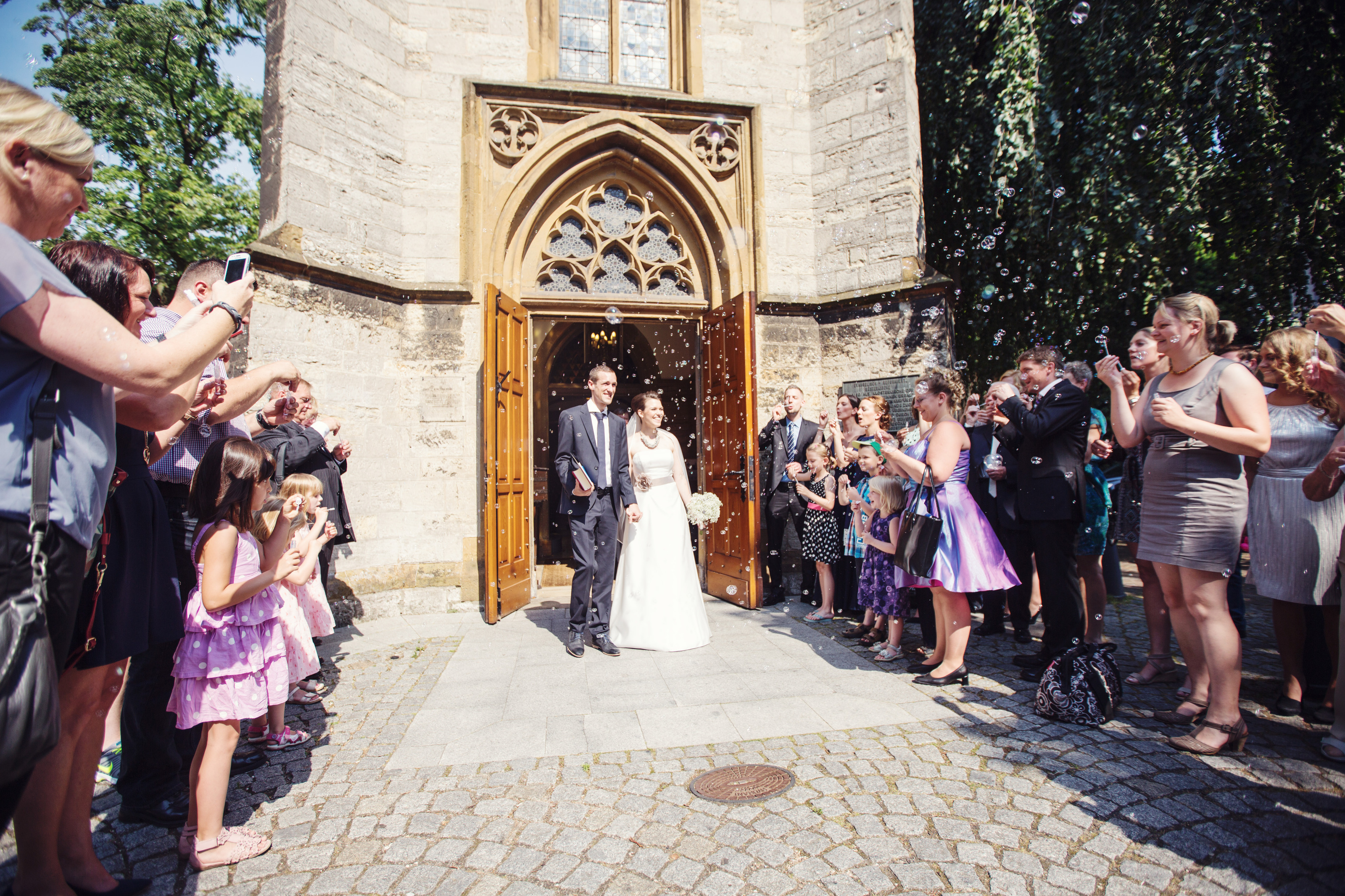 Hochzeitsfotograf Bielefeld – Brautpaar tritt aus gotischem Kirchenportal, jubelnde Gäste beiderseits, Seifenblasen in der Luft
