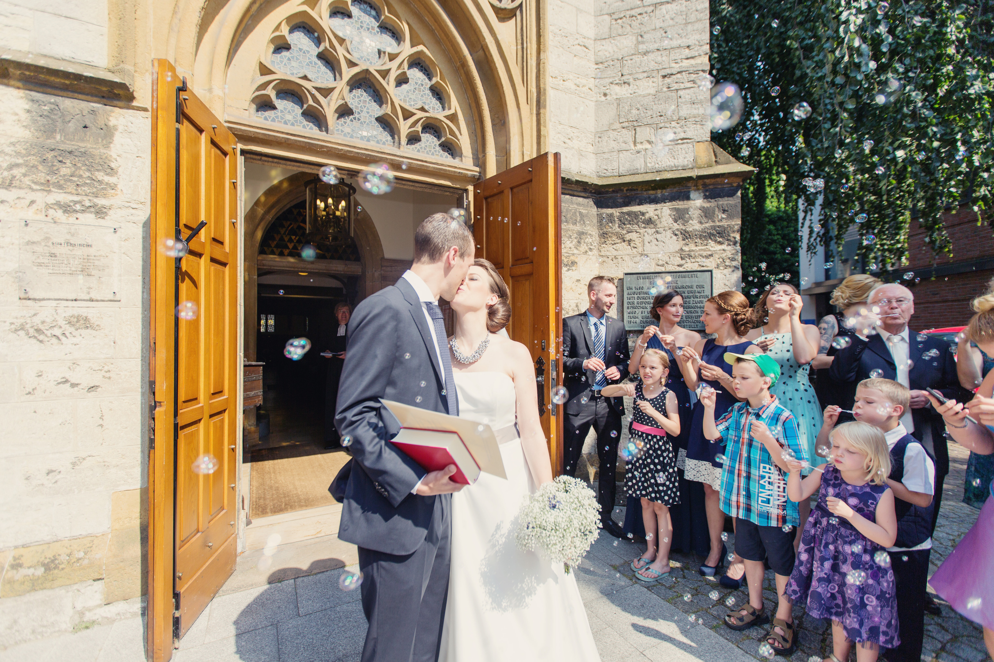 Hochzeitsfotograf Bielefeld – Brautpaar küsst sich vor Kirchenportal, Gäste blasen Seifenblasen, Kinder im Vordergrund