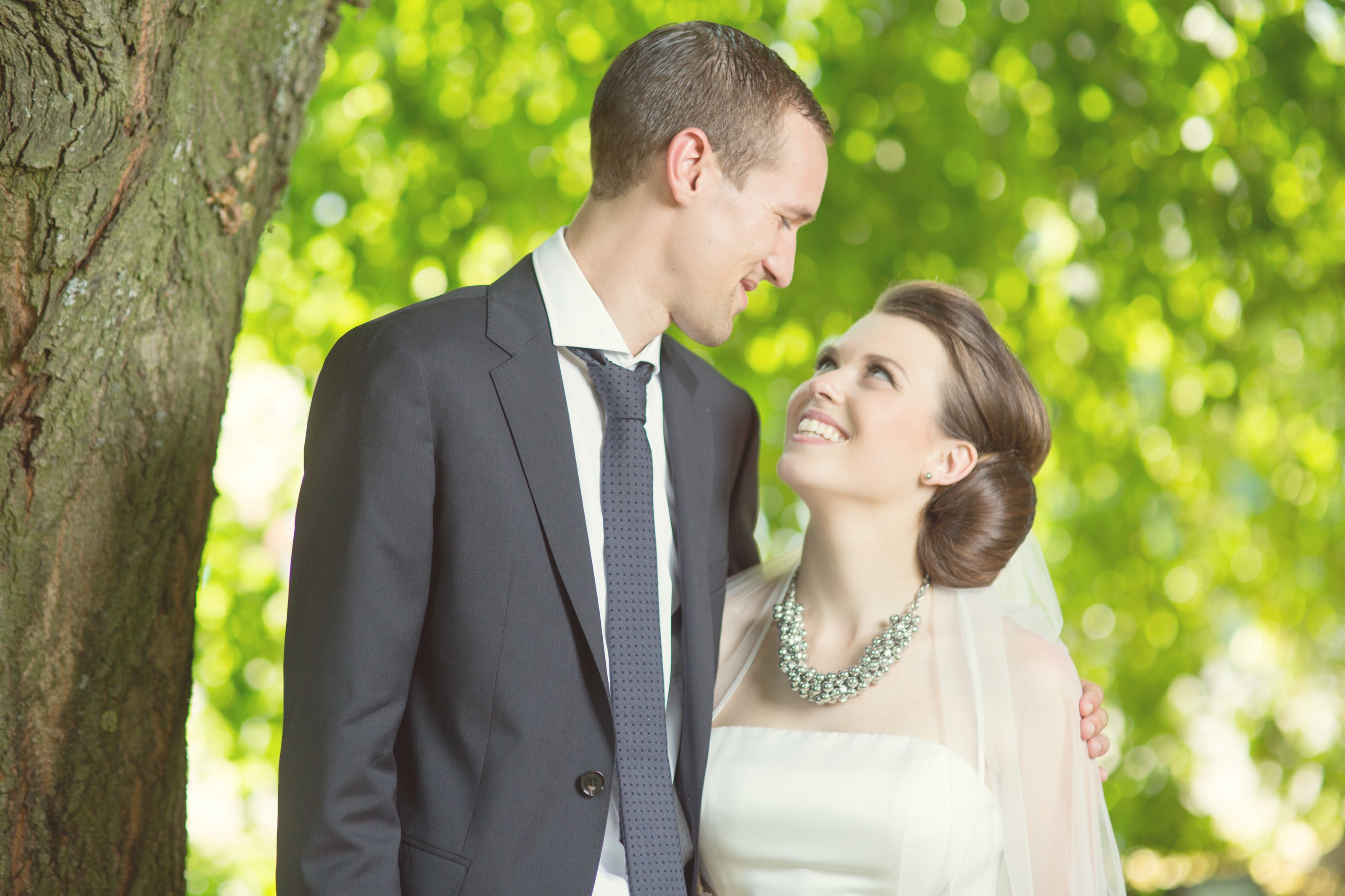Hochzeitsfotograf Bielefeld – Brautpaar steht am Baum, Braut lächelt strahlend den Bräutigam an, grünes Bokeh