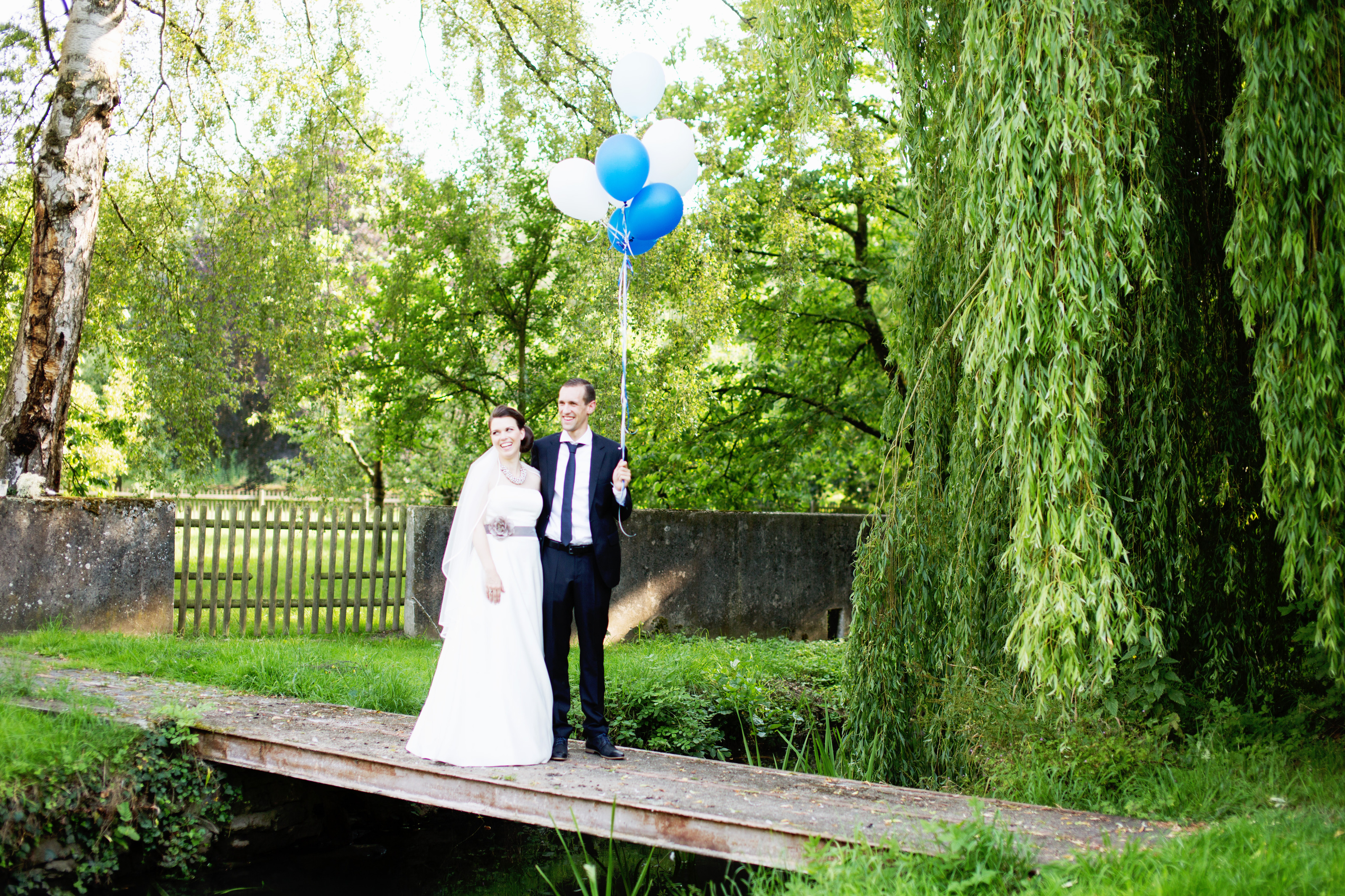 Hochzeitsfotograf Bielefeld – Brautpaar auf Holzbrücke, Bräutigam hält blaue und weiße Luftballons, Trauerweide im Hintergrund