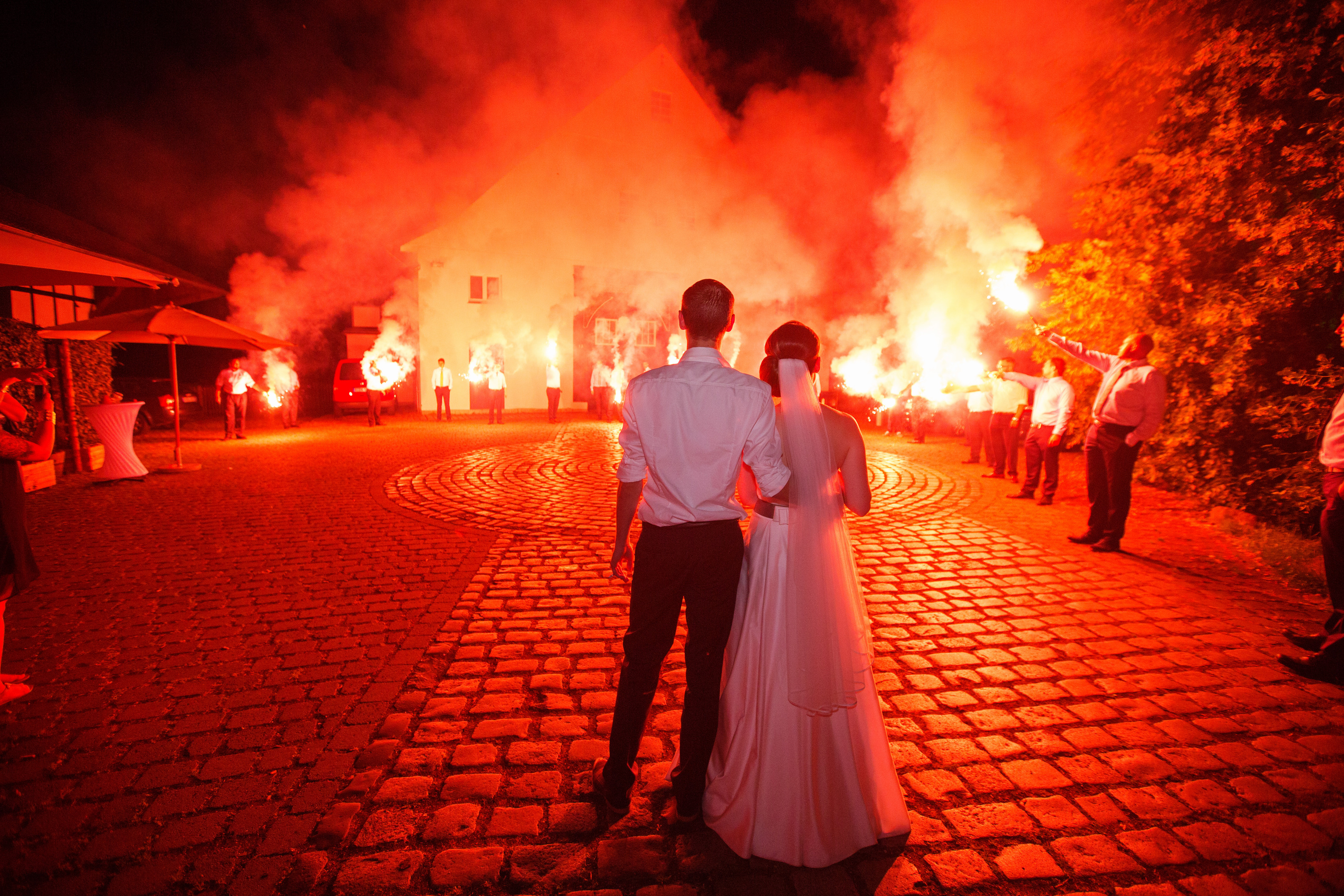 Hochzeitsfotograf Bielefeld – Brautpaar von hinten auf Kopfsteinpflaster, Gäste halten rote Bengalos, dramatische Nachtaufnahme