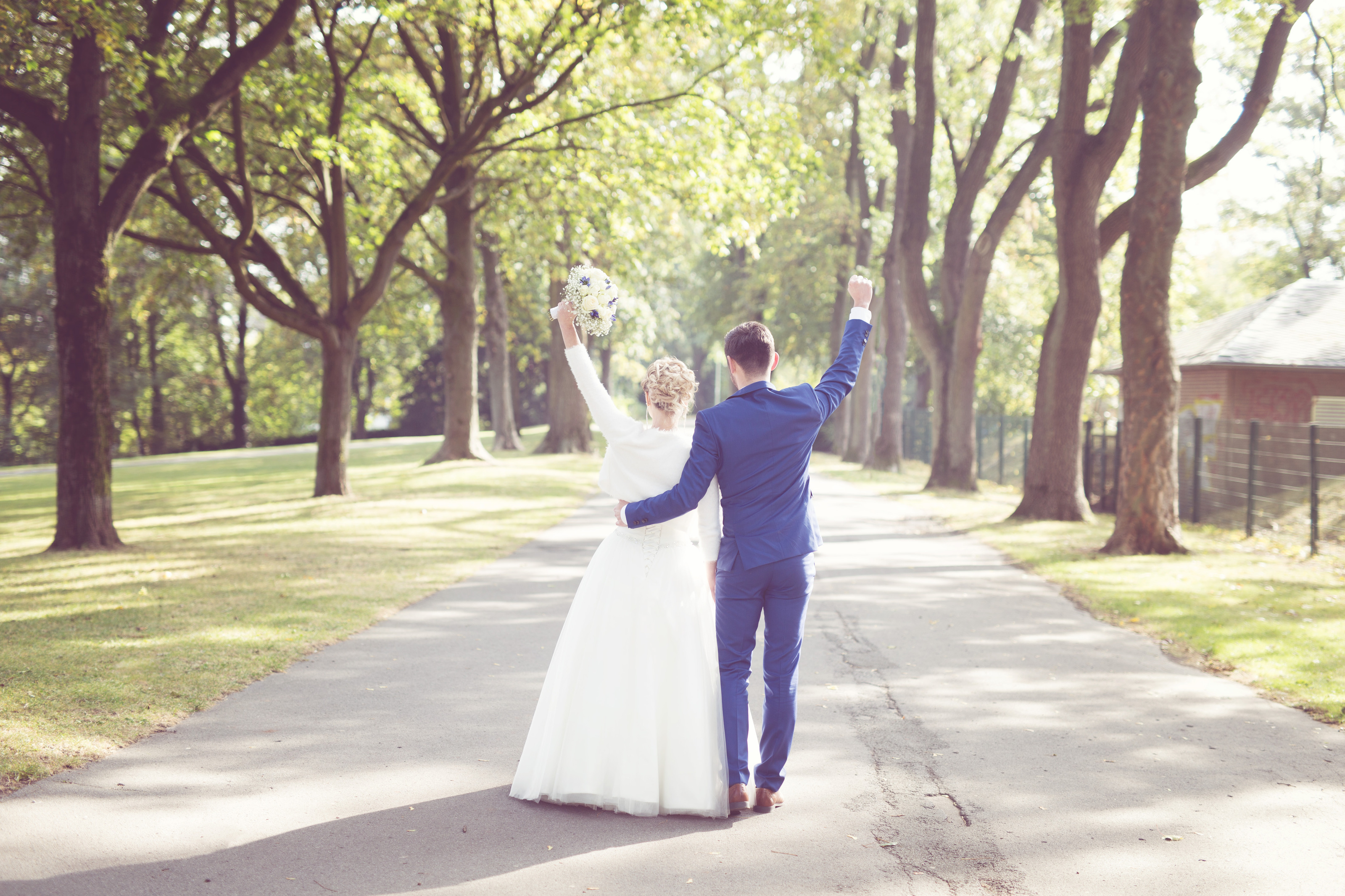 Hochzeitsfotograf Bielefeld – Brautpaar von hinten auf Parkweg, reckt jubelnd die Arme aus, Baumallee, warmes Herbstlicht