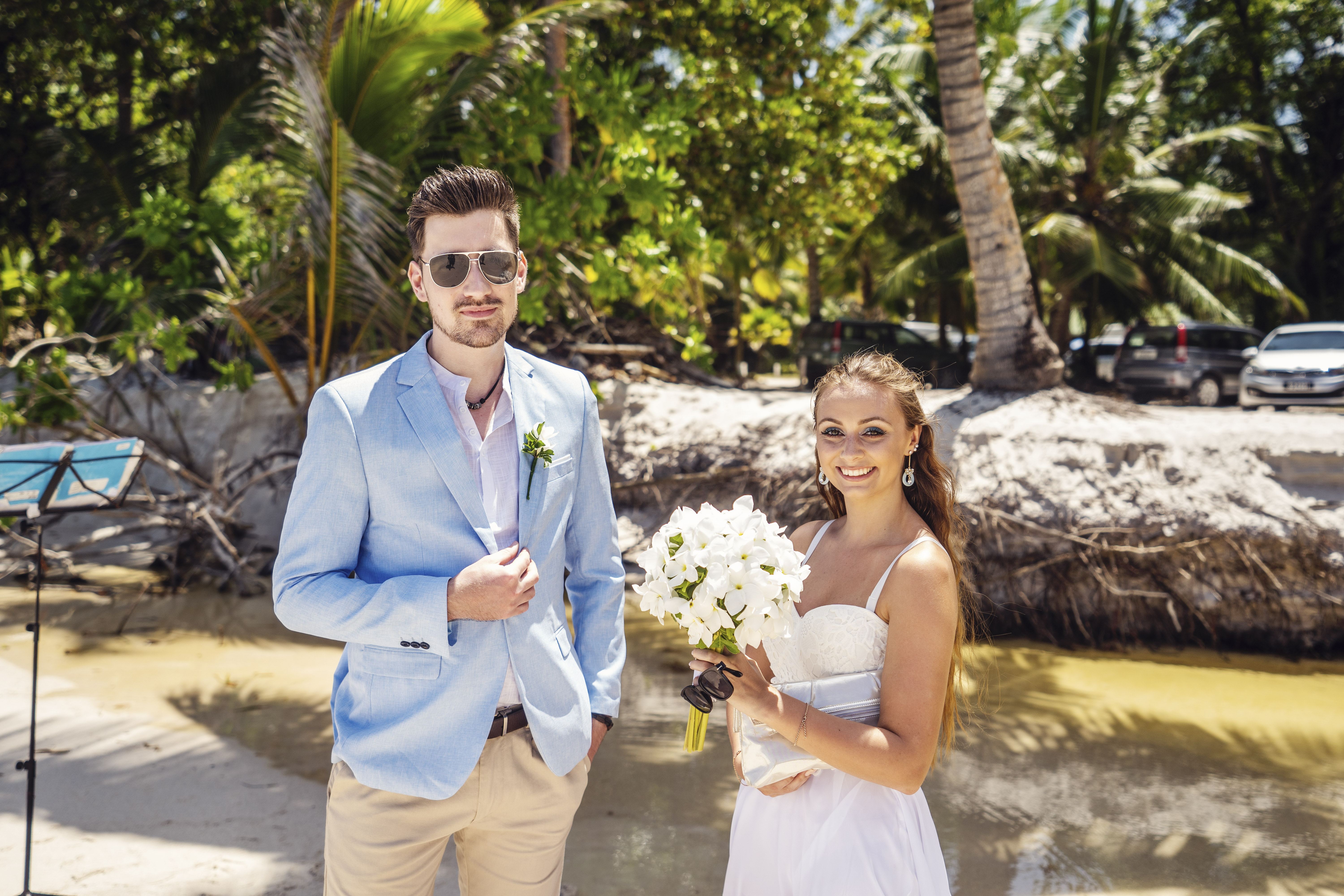 Hochzeitsfotograf Bielefeld – Destination Wedding Seychellen – Brautpaar sitzt auf Felsen am Strand, Braut im weißen Kleid mit Blumenstrauß, Palmen und Meer