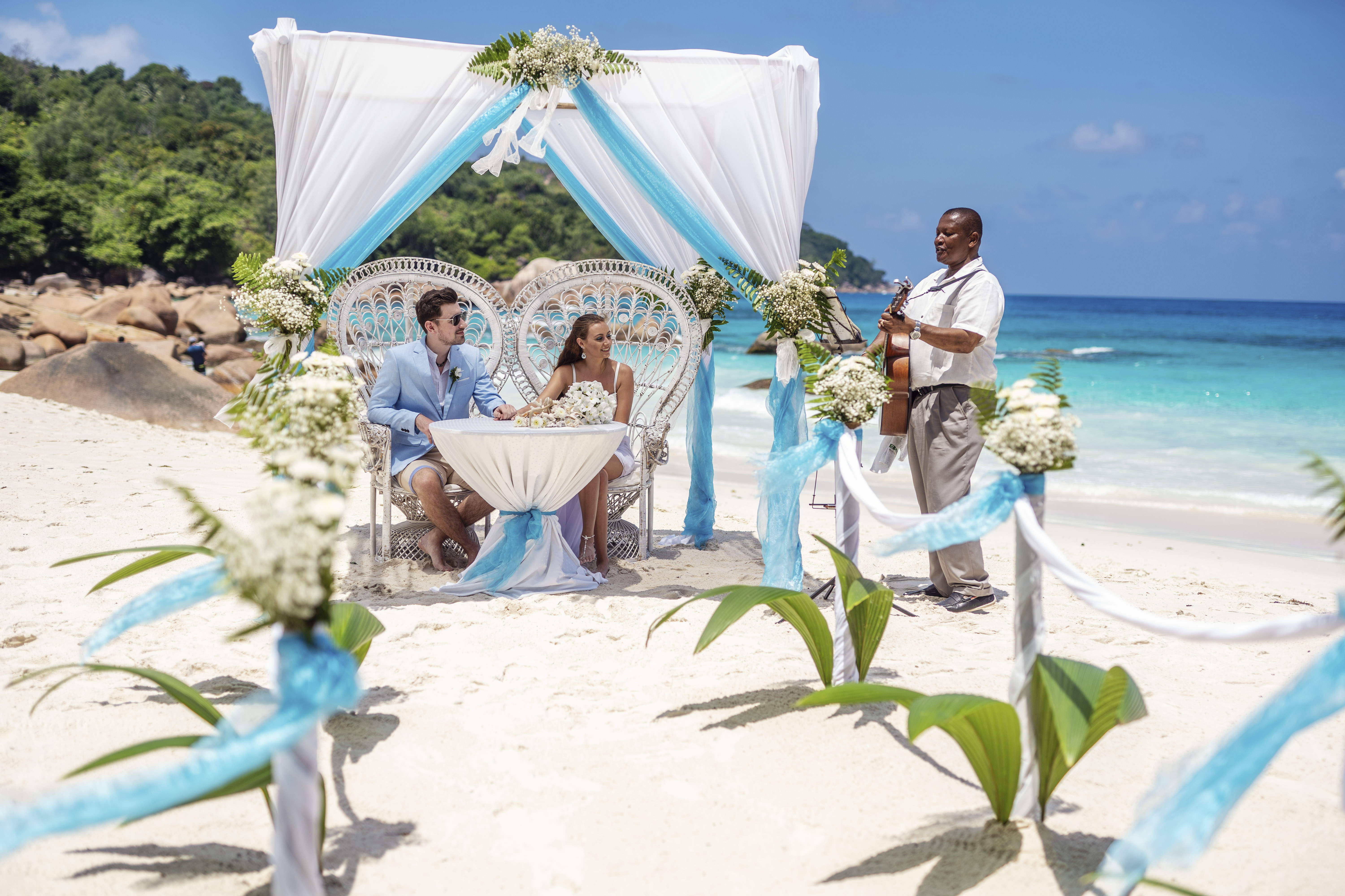 Hochzeitsfotograf Bielefeld – Hochzeitstauung auf den Seychellen unter weißem Baldachin, Brautpaar am Tisch, Musiker mit Gitarre, türkises Meer