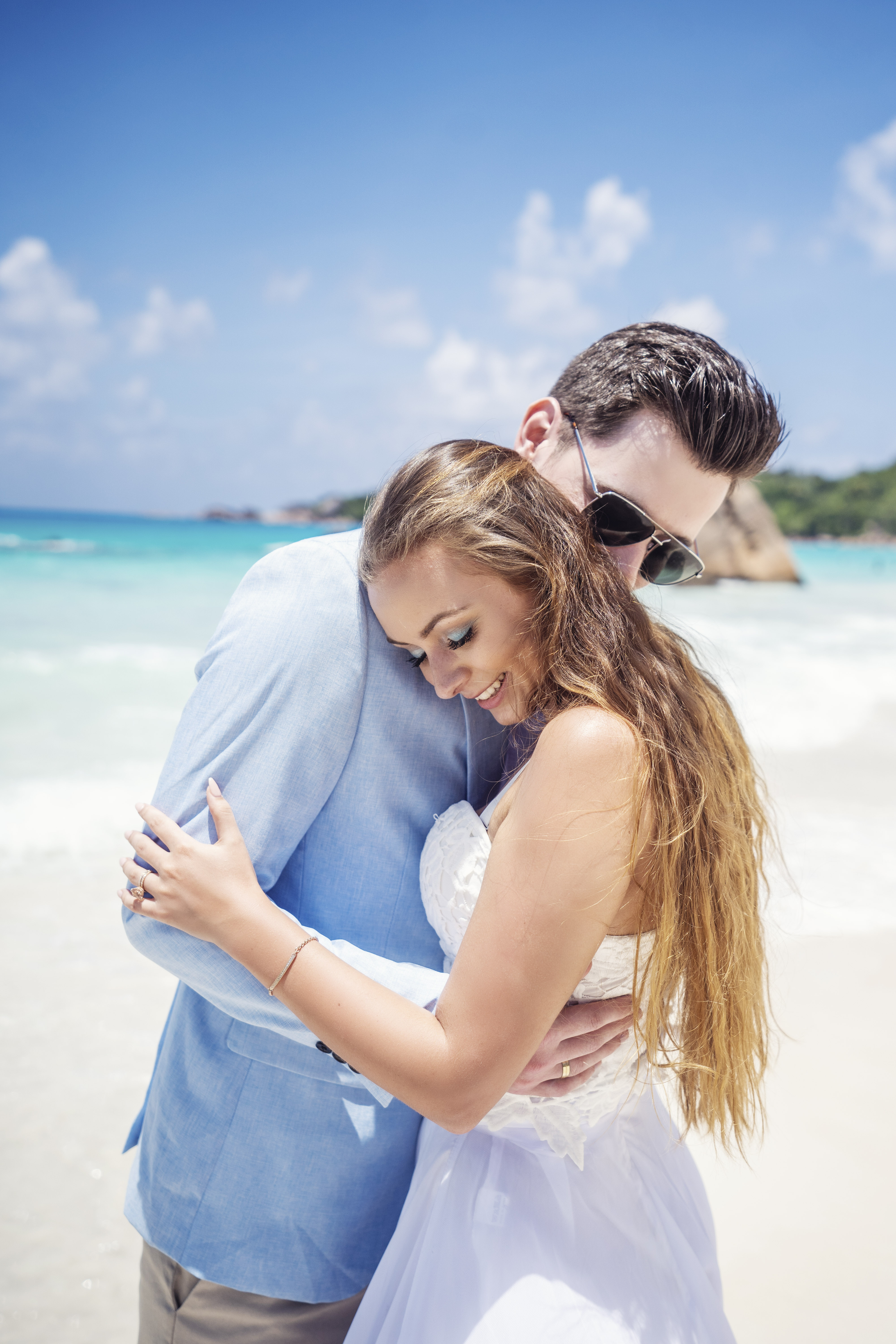 Hochzeitsfotograf Bielefeld – Hochzeitsportrait Seychellen, Brautpaar umarmt sich am Strand, Braut lächelt, Granitfelsen im Hintergrund
