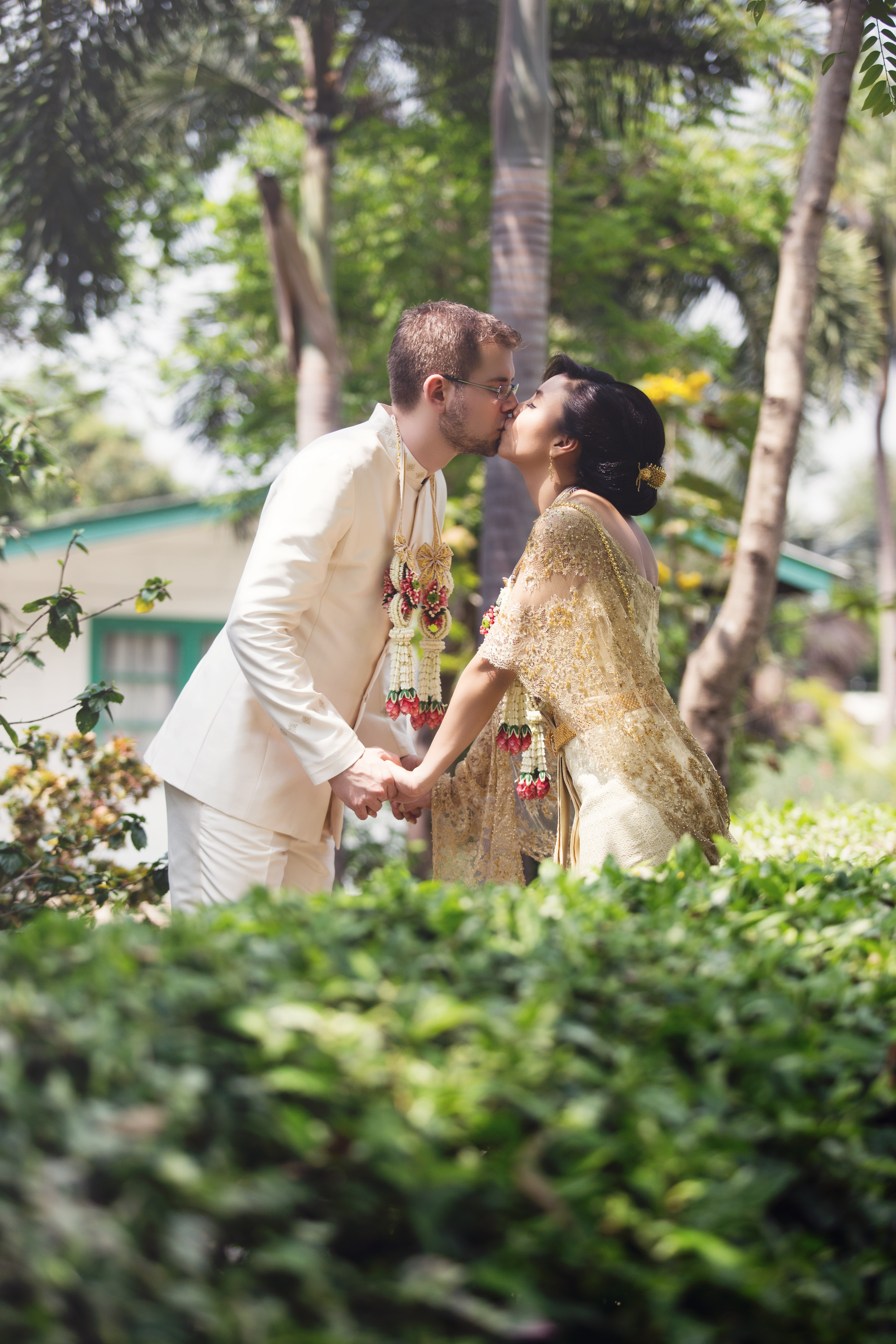 Hochzeitsfotograf Bielefeld – Buddhistische Hochzeit Bangkok, Brautpaar küsst sich in traditioneller Festkleidung im Garten, Blumengirlanden