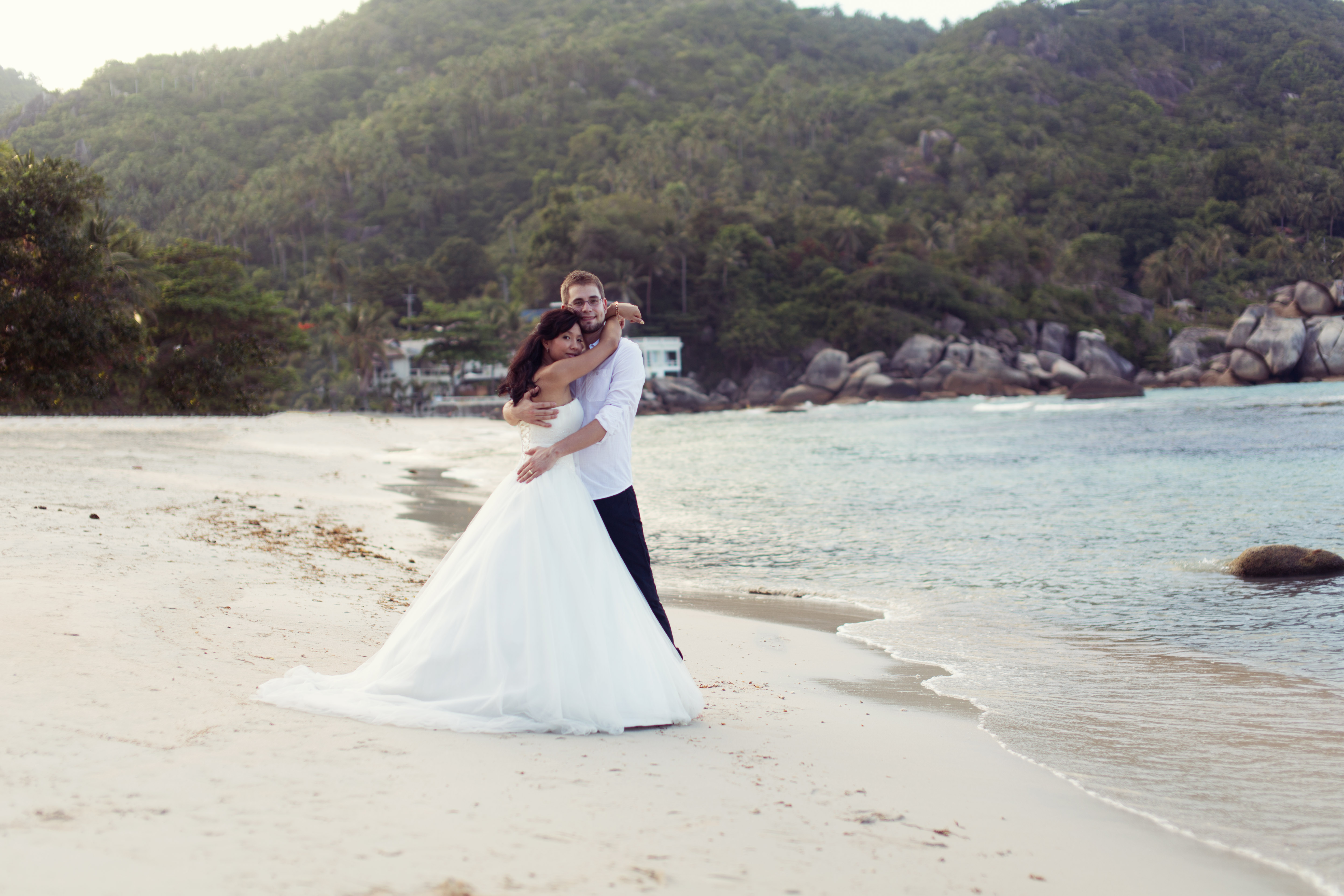 Hochzeitsfotograf Bielefeld – Hochzeitsfotos Thailand, Brautpaar umarmt sich am Strand von Koh Samui, Braut im weißen Kleid, bewaldete Hügel und Meer